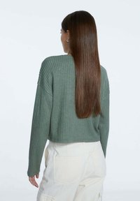 Pull côtelé raccourci en tissu vert pâle avec manches longues, associé à un pantalon de couleur claire. Texture douce et coupe décontractée.