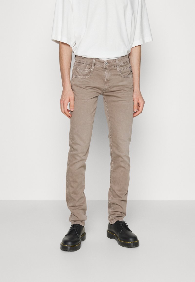 Homme portant un jean beige slim fit et des chaussures noires lacées, debout devant un fond blanc.