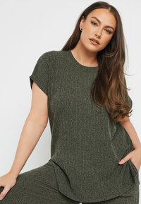 Oliivinvihreä, ribattu neulepaita lyhyillä hihoilla ja pyöreällä kaula-aukolla, jossa on rento malli ja huomaamattomat sivuhalkiot.