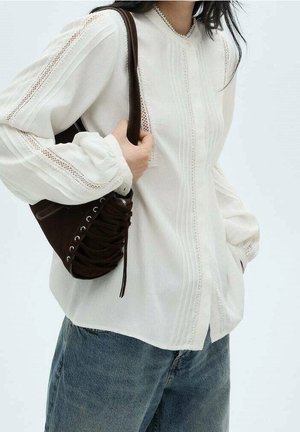 Femme portant un chemisier blanc à manches longues avec des détails en dentelle, un jean bleu, et portant un sac à bandoulière marron foncé avec lacets.