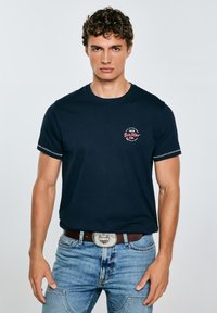 Tumši zils kokvilnas t-krekls ar īsām piedurknēm, uz krūtīm ir sarkans un balts logotips. Valkāts ar gaiši ziliem džinsiem un Sudraba jostu sprādzi.