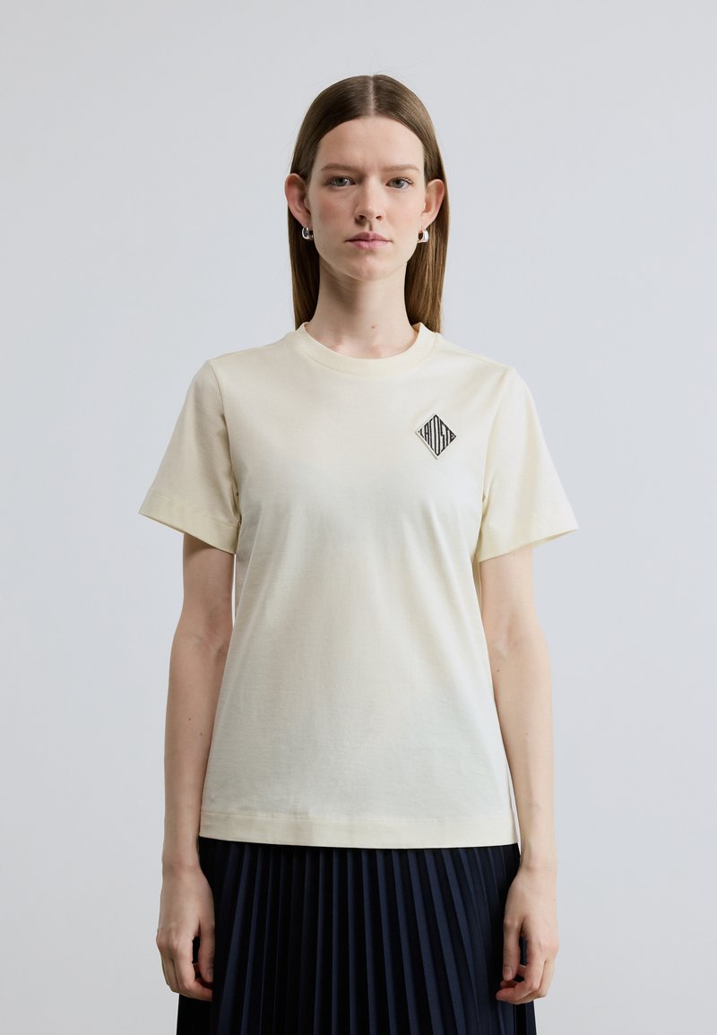 Ανοιχτό κίτρινο βαμβακερό T-shirt με κοντά μανίκια, στρογγυλή λαιμόκοψη και μαύρο γραφικό σε σχήμα ρόμβου στο αριστερό μέρος του στήθους. Ομαλή υφή.