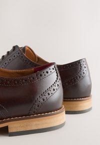 Paire de chaussures habillées brogues en cuir marron foncé avec détails perforés et talons empilés en bois sur fond clair.