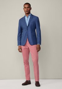 Man i blå blazer, ljusblå skjorta, rosa byxor och bruna loafers som står mot en beige bakgrund.