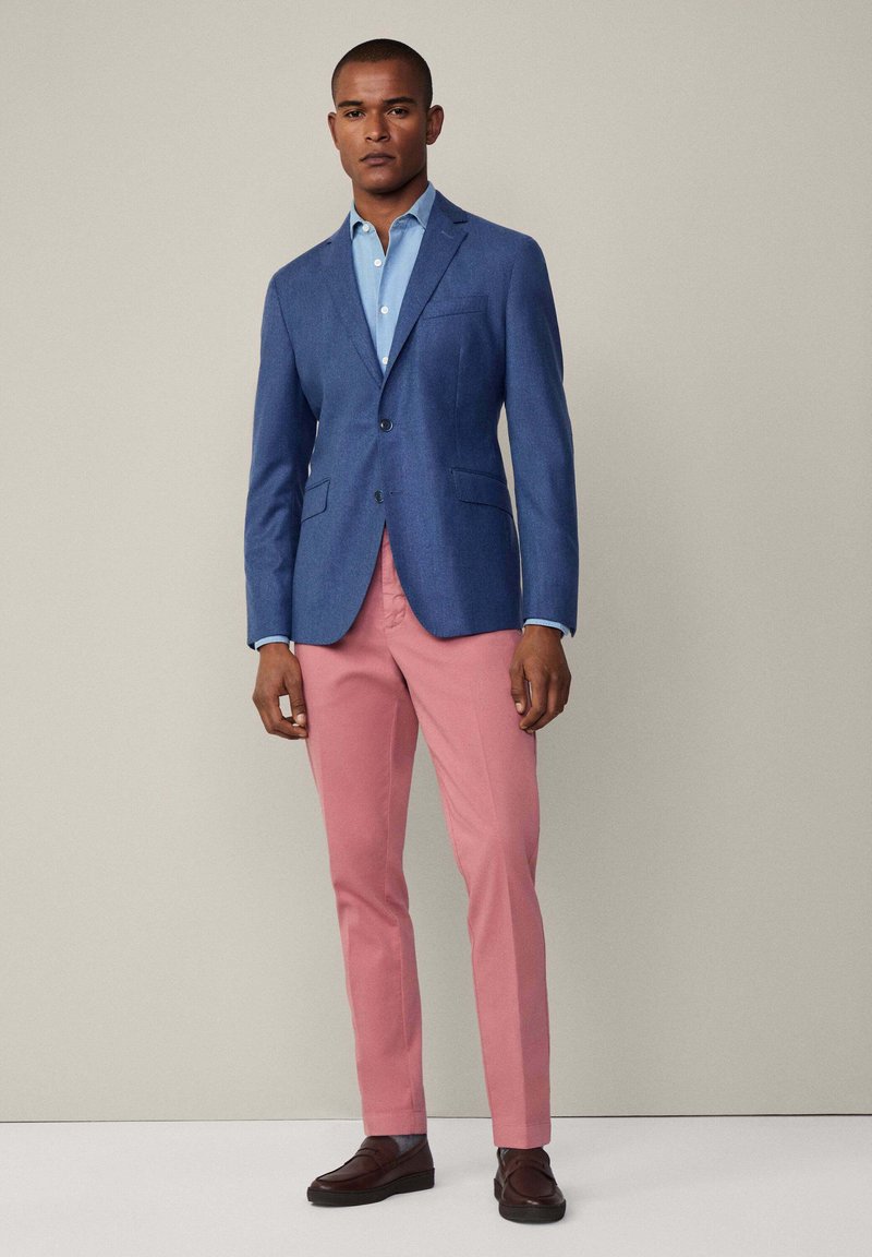 Man in een blauwe blazer, lichtblauw overhemd, roze broek en bruine loafers staat tegen een beige achtergrond.