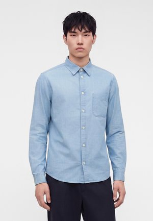 Jeune homme portant une chemise bleu clair à boutons avec une poche poitrine et un pantalon foncé, debout devant un fond blanc uni.