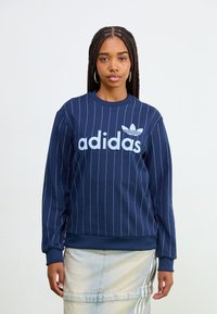 Mörkblå sweatshirt med vertikala vita ränder, med en stor ljusblå "adidas"-logotyp och logotype emblem på bröstet, ribbade muddar och nederkant.