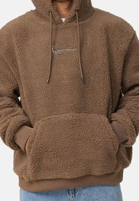 Brun sherpa-fleece hoodie med framficka och broderad logotyp på bröstet, bärs med ljusblå jeans.