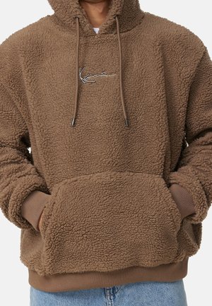 Brown sherpa fleece hoodie with front pouch pocket and embroidered logo on chest, worn with light blue jeans.