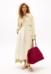 Cappotto trench beige doppiopetto con bottoni dorati, abito bianco, borsa a tracolla rosa intrecciata e scarpe beige slip-on con finitura testurizzata.