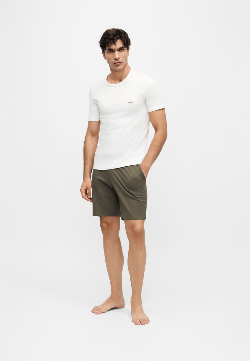 Homme debout pieds nus portant une chemise blanche à manches courtes et un short vert olive avec une main dans la poche sur fond uni.