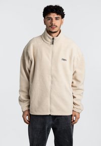 Giacca in pile color crema con colletto alto, zip frontale e dettaglio del logo sul lato sinistro del petto. Presenta maniche lunghe e una vestibilità rilassata.