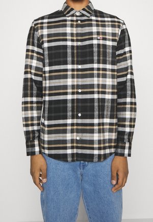 Personne portant une chemise à carreaux noire, grise, blanche et beige avec boutons et manches longues, ainsi qu’un jean bleu, debout devant un fond uni.