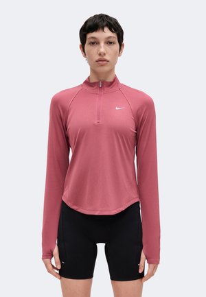 Ung person med kort mørkt hår iført en pink langærmet træningstrøje og sorte cykelshorts, stående mod en ensfarvet baggrund.