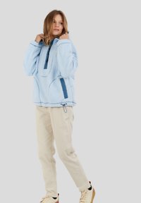 Vrouw in een lichtblauwe fleece trui, beige broek en crème sneakers, staande tegen een effen grijze achtergrond.