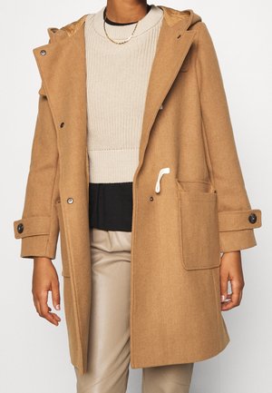 Personne portant un manteau en laine couleur camel avec de grandes poches, des poignets boutonnés et un pull en tricot beige en dessous.