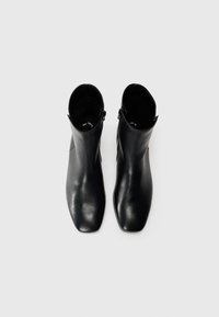 Par de botas de cuero negro hasta el tobillo con puntas cuadradas y cremalleras laterales, vistas desde arriba sobre un fondo gris claro.