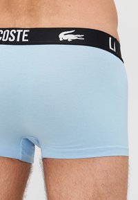 Svijetloplave pamučne bokserice s crnim pojasom na kojem se nalazi bijelo "LACOSTE" brendiranje i logotip. Tkanina je meka i glatka.