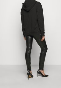 Sudadera negra con capucha y pantalones de cuero negro ajustados, con textura brillante y tacones de aguja negros puntiagudos, vistos desde atrás.