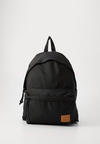 Sac à dos noir avec une texture de tissu lisse, un dessus arrondi, une poche zippée à l'avant, des bretelles réglables et un patch en cuir marron.
