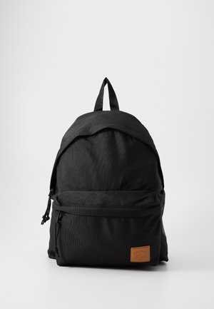 Mochila negra con una textura de tela suave, parte superior redondeada, bolsillo frontal con cremallera, correas ajustables y un parche de cuero marrón con el logo.