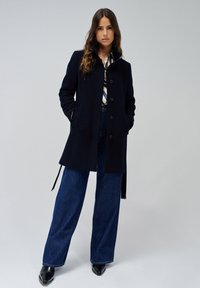Manteau marine foncé avec col et fermeture à boutons, doté de poches latérales. Associé à un denim bleu à jambes larges et des chaussures noires.