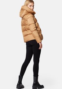 Kurtka puchowa w jasnobrązowym kolorze z kapturem, z pikowanym wzorem i przednimi kieszeniami, w zestawieniu z dopasowanymi czarnymi spodniami i masywnymi czarnymi butami.