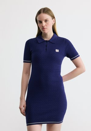 Kobieta ubrana w granatową sukienkę polo z krótkim rękawem i białym obszyciem, wykonanej z dzianiny warkoczowej, stojąca na tle jasnego, jednolitego tła.