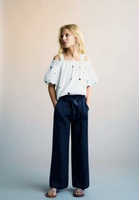 Blusa bianca off-shoulder con pois blu, abbinata a pantaloni a gamba larga blu scuro e sandali con fasce, con un fiocco in vita.