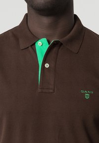 Polo marron avec un plastron de col vert, deux boutons et un logo "GANT" brodé en vert sur la poitrine. Texture douce en coton.