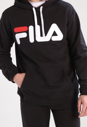 Person wearing a black FILA hoodie with white and red logo, hands in front pocket, paired with black pants against a plain background.