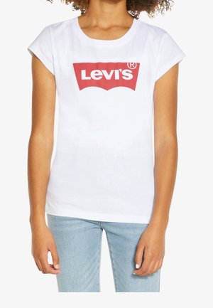 Kobieta ubrana w białą koszulkę Levi's z czerwonym logo i jasnoniebieskie dżinsy, stojąca z rękami swobodnie opuszczonymi wzdłuż boków.