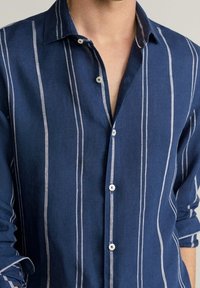 Modrá pruhovaná lněná košile s límcem, zapínáním na jeden knoflík a vykasanými rukávy. Světle šedé vertikální pruhy kontrastují s tmavě modrou tkaninou.