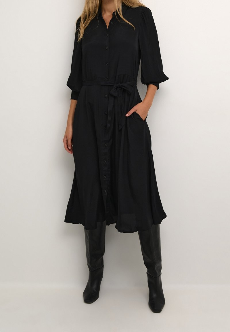 Robe noire boutonnée avec manches trois-quarts, ceinture à la taille et poches latérales. Confectionnée dans un tissu lisse et léger.