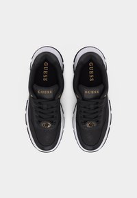 Scarpe da ginnastica Guess nere con suole bianche, emblema dorato a forma di "G" sui lacci e logo Guess dorato sulla linguetta viste dall'alto.