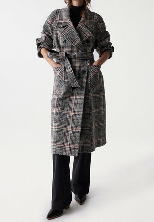 Femme portant un long manteau à carreaux noir, gris et rouge avec ceinture, sur un pantalon noir à jambes larges et des chaussures noires pointues, les mains dans les poches du manteau.