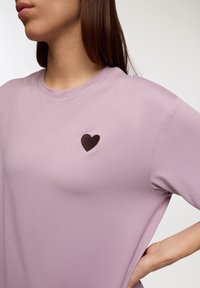 Maglietta a maniche corte lavanda con un cuore ricamato marrone scuro. Tessuto liscio, scollatura a girocollo e vestibilità rilassata.