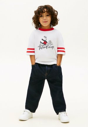 Enfant aux cheveux bouclés se tient les mains dans les poches, portant une chemise blanche à rayures rouges, un jean bleu et des baskets blanches.