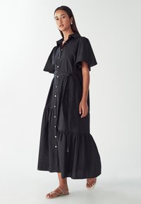 Čierne bavlnené maxi šaty s krátkymi volánovými rukávmi, gombíkovým zapínaním vpredu, opaskom v páse a sukňou v úrovniach. Nosené s plochými sandálmi na remienky.