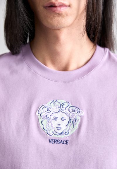 Lavendel värvi puuvillane t-särk, millel on keerukate detailidega tikand Medusa kujutisega ja sõna "VERSACE" graafika all.