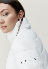 Chaqueta de plumas blanca con cuello alto, textura acolchada y detalle del logo negro en la manga. El modelo lleva el pelo largo y pendientes de botón.
