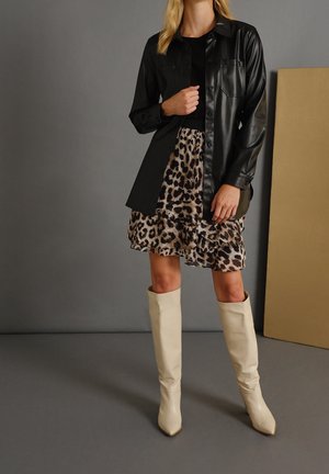 Donna che indossa un giubbotto di pelle nera con bottoni, una gonna con stampa leopardata e stivali beige al ginocchio, in piedi davanti a uno sfondo grigio e beige.