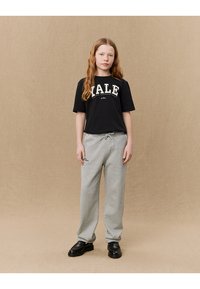 Schwarzes Baumwoll-T-Shirt mit "YALE" Aufdruck, kombiniert mit hellgrauen Jogginghosen und schwarzen Slippern. Vor neutralem Hintergrund.