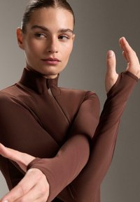 Top lung maro cu mâneci lungi, guler înalt și găuri pentru degete, fabricat dintr-un material neted și elastic. Modelul pozează cu brațele încrucișate, mâinile întinse.