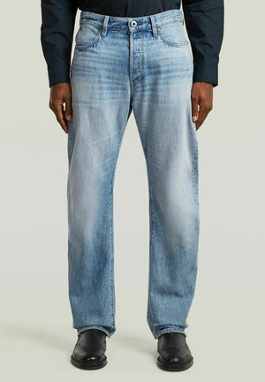 Mand iført lyseblå jeans med lige ben, sorte sko og en sort langærmet skjorte, stående foran en ensfarvet baggrund.