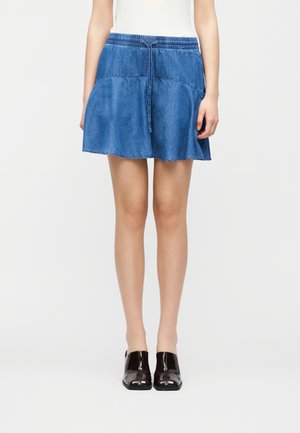 Femme portant une jupe en denim bleu longueur genou avec une taille à cordon de serrage et des chaussures noires brillantes à bout carré, debout devant un fond blanc uni.