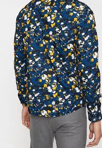 Blus med lång ärm i blått tyg med ett flerfärgat blommönster. Har en spetsig krage och en avslappnad passform, med knapppdetaljer.