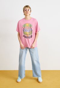Pink oversized t-shirt with a beach scene graphic, light blue wide-leg jeans, and mint green sneakers, paired with pink sunglasses.