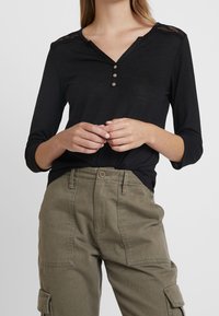 Svart långärmad topp med V-ringning, knappslå och spetsdetaljer på axlarna; matchad med olivgröna cargobyxor med fickor.