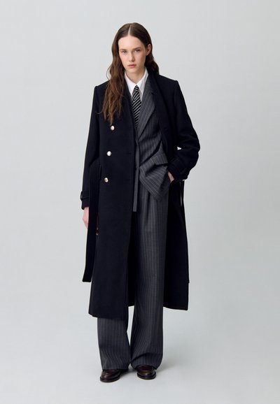 Cappotto doppiopetto nero con bottoni dorati, indossato sopra pantaloni e giacca a righe, completato con una camicia bianca e una cravatta a righe.